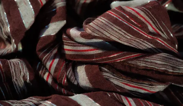 Görüntü. İnce kırmızı aksanlı ve metalik parlayan kahverengi ve beyaz çizgili bir kumaş. Kumaşın kıvrımları ve dokusu zengin ve dinamik bir görüntü yaratır.