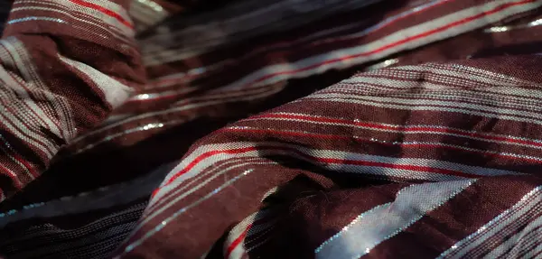 Beyaz ve kırmızı zıt çizgilerle süslenmiş kestane rengi kumaşın detaylı bir görüntüsü. Gümüş ipliklerin ince parıltısı eşsiz, pırıl pırıl bir etki yaratarak zengin renk paletini vurguluyor.