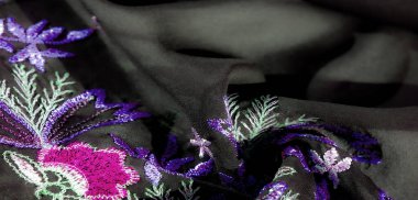 Koyu renk kumaş üzerine ayrıntılı nakış bulanıklığı, yeşil aksanlı mor ve pembe çiçek desenlerinin zengin bir kombinasyonunu gösteriyor. Katlama ve dikişler sanatsal bir derinlik yaratır..