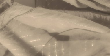 Yumuşak ışıkta hafifçe parıldayan yatay çizgileri olan açık renkli kumaş bulanıklığı. Katmanlı dokular ve katlar derinlik ve incelik ekler..