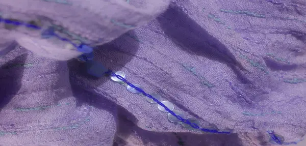 Hassas pullarla süslenmiş soluk mor bir kumaş. Yeşil ve mavi dikişli çizgiler boyunca dizilmiş. Yumuşak, katlanmış yüzeylerde ince bir doku oluşturuyor..