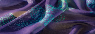 Işıldayan turkuaz desenlerle süslenmiş, yumuşak dokular ve gölgeler yaratmak için nazikçe katlanmış mor bir kumaşın yakın çekimi. Karmaşık detaylar ve canlı renkler uyum içinde harmanlanır.