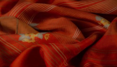 Renkli çiçek desenleri ve beyaz çizgili aksanlı turuncu kumaşın bulanık görüntüsü. Yumuşak, akışkan kıvrımlar sıcaklık ve sanatsal tasarım hissi yaratıyor..