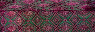 Karmaşık yeşil ve pembe geometrik desenli bulanık bir kumaş. Kumaş, canlı renkler arasında keskin bir kontrastla zengin bir doku oluşturan ayrıntılı bir nakış tasarımı içerir..