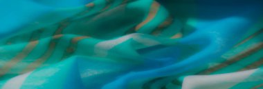 Bulanık mavi ve yeşil kumaş yumuşak dalgalarda altın çizgilerle vurgulanır ve modern tekstil sanatını yansıtan zengin bir doku yaratır..