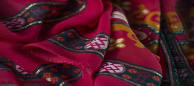 Bulanık kırmızı kumaşın zengin dokusunu parlak geometrik desenler, turuncu çiçekli motifler ve parıldayan yeşil detaylarla vurgular.