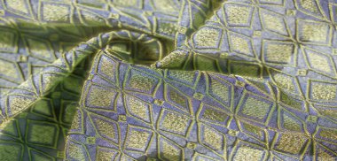 Bu kumaşın karmaşık geometrik desenlerini bulanıklaştırarak, dokulu mavi tabana karşı yeşil ve altın renklerini sergiliyor, lüks ve sanatsal bir etki yaratıyor..