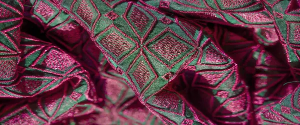 Pembe ve yeşil tonlarda, pürüzsüz katları ve geometrik nakışları olan lüks bir kumaşın bulanık fotoğrafı. Karmaşık desenler ve canlı renkler zengin ve zarif bir görünüm yaratır..