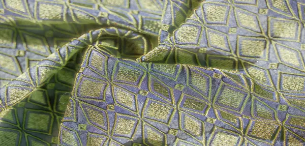 Bu kumaşın karmaşık geometrik desenlerini bulanıklaştırarak, dokulu mavi tabana karşı yeşil ve altın renklerini sergiliyor, lüks ve sanatsal bir etki yaratıyor..