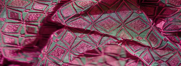 Pembe ve yeşil renkli, karmaşık desenli, canlı, nakışlı kumaşın bulanık yakın plan görüntüsü. Geometrik şekiller ve detaylı dikişler göz alıcı bir görsel doku oluşturuyor..