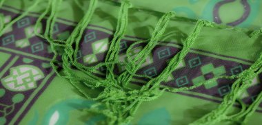 Karmaşık mor desenleri ve püsküllü kenarları olan canlı yeşil bir kumaş dokuda dinamik bir renk ve doku oyunu yaratır. Cesur tasarımlar zengin bir estetik cazibe ekler..