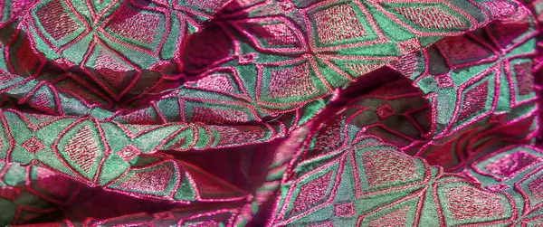 Pembe ve yeşilin canlı tonlarında geometrik desenli, dokulu bir kumaşın bulanık görüntüsü. Karmaşık nakış zengin ayrıntıları gösteriyor, lüks bir görünüm yaratıyor..