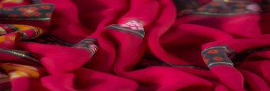 Karmaşık turuncu, beyaz ve sarı çiçek desenleri ve parıltılı aksanlarıyla canlı kırmızı kumaşın bulanık görüntüsü, sıcak ve renkli bir tasarım sergiliyor..