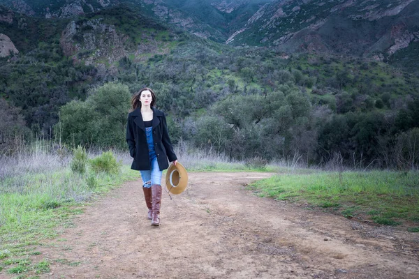 Country girl dressed in bib overalls, black jacket and boots walking on a dirt road with mountain in the background.