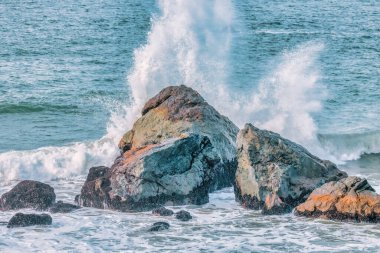 Su sıçrıyor. Kristal berrak deniz suyu kayalara çarpıyor. Deniz dalgaları okyanusun taştan kıyılarında kırılır, mavi su pastel renklerde.