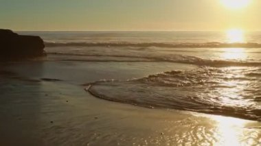 Kumsalda altın bir gün batımı, köpüklü dalgalar beyaz kumsala doğru yuvarlanıyor, güneş ışığı altında.