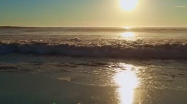 Kumsalda altın gün batımı, köpüklü dalgalar beyaz kumsala doğru yuvarlanıyor güneş ışığı altında