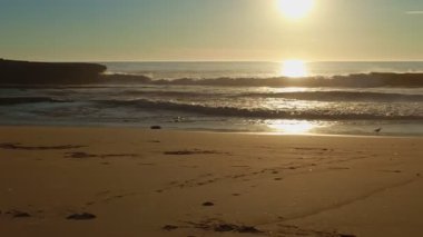 Kumsalda altın bir gün batımı, köpüklü dalgalar beyaz kumsala doğru yuvarlanıyor, güneş ışığı altında.