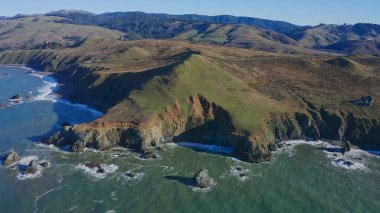 1. Otoyol 'daki engebeli Kaliforniya kıyı şeridinin nefes kesici manzarası. Dalgaların kayalık oluşumlara çarpmasıyla derin mavi okyanusun üzerinde yeşil bitki örtüsüyle kaplı dik uçurumlar yükselir. Dönek sahil yolu çarpıcı manzaralı bir yol sunuyor.