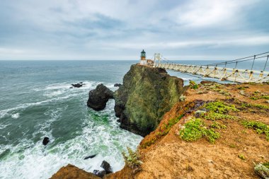 San Francisco, Kaliforniya yakınlarındaki Point Bonita Deniz Feneri Pasifik Okyanusu üzerindeki engebeli bir uçurumun üzerinde duruyor. Üzerinde paslı bir fener kulesi ve bulutlu bir gökyüzünün altındaki tarihi asma köprü bulunuyor.