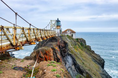San Francisco, Kaliforniya yakınlarındaki Point Bonita Deniz Feneri Pasifik Okyanusu üzerindeki engebeli bir uçurumun üzerinde duruyor. Üzerinde paslı bir fener kulesi ve bulutlu bir gökyüzünün altındaki tarihi asma köprü bulunuyor.