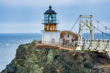 San Francisco, Kaliforniya yakınlarındaki Point Bonita Deniz Feneri Pasifik Okyanusu üzerindeki engebeli bir uçurumun üzerinde duruyor. Üzerinde paslı bir fener kulesi ve bulutlu bir gökyüzünün altındaki tarihi asma köprü bulunuyor.