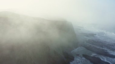 Top view of foggy coastal cliffs in California. Ocean waves crash against the rocks below as thick mist rolls over the shoreline. Moody and dramatic nature footage perfect for travel and cinematic themes
