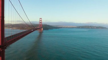 Golden Gate Köprüsü, gündoğumunda arka planda San Francisco şehir merkezi silueti olacak. Yumuşak sabah ışığında ikonik Kaliforniya manzarası, huzurlu okyanus ve berrak gökyüzü.