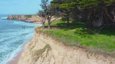 Davenport, California 'daki sahil uçurumlarının ve kumlu plajların havadan görüntüsü. Okyanus dalgaları engebeli kaya oluşumları ve selvi ağaçlarının yakınındaki kıyılara doğru akar. Seyahat, doğa ve kıyı müziği için mükemmel.