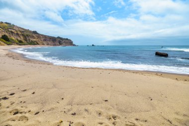 Kaliforniya Pasifik Sahili 'ndeki Greyhound Rock Sahili' nde altın kum ve kayalık sahil şeridi. 1. Otoyol boyunca, parçalı bulutlu bir gökyüzü altında dramatik uçurumlar ve turkuaz dalgalarla manzaralı bir okyanus manzarası.