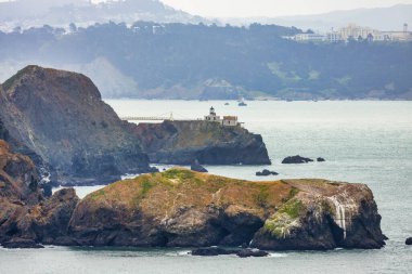 Point Bonita Deniz Feneri 'nin manzarası Marin Burunları, Kaliforniya' da kayalık bir uçuruma tünemişti. Arka planda Pasifik Okyanusu ve sisli tepeler bulunan Rodeo Sahili Sahil Yolu 'ndan çekilmişti.
