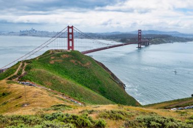 San Francisco Körfezi 'ni kaplayan Golden Gate Köprüsü' nün panoramik manzarası. Şehir manzarası, yemyeşil tepeler ve tekneler bulutlu bir bahar gününde yüksek bir noktadan çekiliyor.