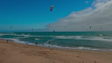 Kaliforniya, ABD - 24 Mayıs 2025, uçurtmacılar 1. Otoyol boyunca Pacifica, California yakınlarında okyanus dalgalarına biniyorlar. Renkli uçurtmalar bu hareketli kıyı sahnesinde turkuaz denizin üzerindeki bulutlu gökyüzünü dolduruyor.