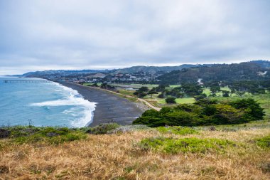 Pasifik, Kaliforniya 'daki Mori Point' ten geniş bir manzara. Çimenli kayalık Pasifik Okyanusu 'na ve bulutlu gökyüzünün altındaki uzak Pacifica İskelesine bakar ve kıyı şeridinin engebeli güzelliğini yakalar.