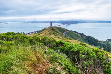 San Francisco Körfezi 'ni kaplayan Golden Gate Köprüsü' nün panoramik manzarası. Şehir manzarası, yemyeşil tepeler ve tekneler bulutlu bir bahar gününde yüksek bir noktadan çekiliyor.