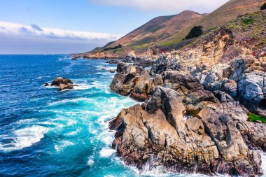 Rocky shoreline along Highway 1 in Big Sur, California, USA. Rugged cliffs meet turquoise Pacific waves, with coastal mountains rising in the background, showcasing iconic scenery of the Pacific Coast Highway.