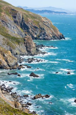 Steep cliffs along the Pacific Ocean near Muir Beach Cove, viewed from Tennessee Valley Trail in Marin Headlands, California. Rugged coastline with waves crashing on rocks under a bright sky