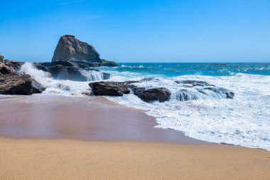 Scenic sandy beach along Highway 1 California with ocean waves crashing on rocks under clear blue sky and coastal cliffs capturing the beauty of the Pacific Coast on a sunny summer day