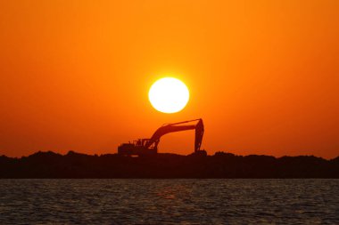 Gün batımında yat limanı inşaatı için kayaların üzerine kazıcı silueti yerleştirilmiş. Güneş ışıl ışıl parlarken.