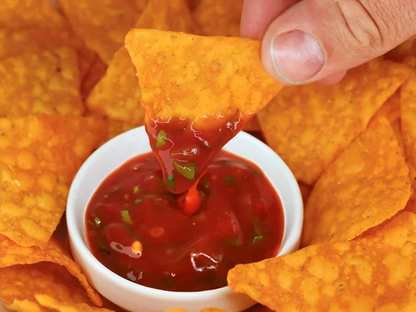 close up of dipping a tortilla chip in hot spicy salsa with chili peppers, classic mexican corn chips as snack for partys served in a bowl.