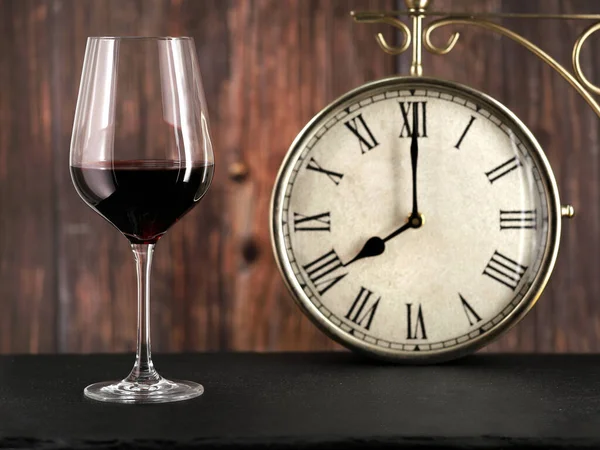 Glass of red wine with antique clock on wooden background, concept image for dinner time or alcoholism in the evening after work.