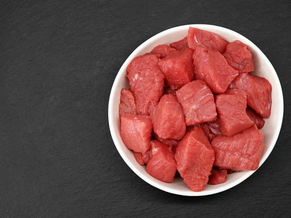raw diced beef meat in white bowl on black slate background, top view.