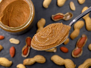 Kavanozlu bir kaşık kremalı fıstık ezmesi ve koyu arkaplanda kabuklu fıstık..