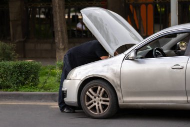 Bir adam yolda bozuk arabasını tamir etmeye çalışıyor, kaput açık.