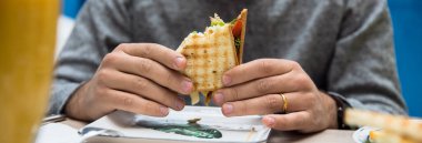 Adam kafede oturmuş sandviç yiyor.