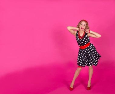 Siyah elbiseli, kırmızı aksesuarlı, beyaz benekli, pembe elbiseli sarışın bir kız: yüksek topuklu ayakkabılar, bilezik ve küpeli boncuklar, kafasında halka. Kırmızı rujlu dudaklar. Pin-up.