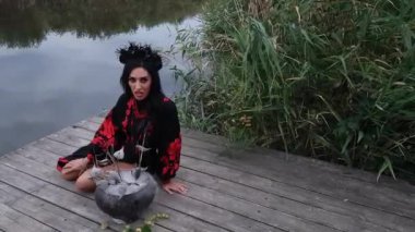 A girl in an embroidered black and red dress with beads, in a black wreath, cooks with herbs in a cast-iron cauldron and conjures, sitting on a pier on the river bank against the background of reeds
