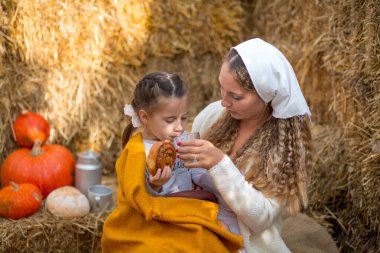 Annesi kızına tarçınlı börek yediriyor ve arka plandaki saman, balkabağı, ekmek ve fincanla birlikte içeceği sütü veriyor.