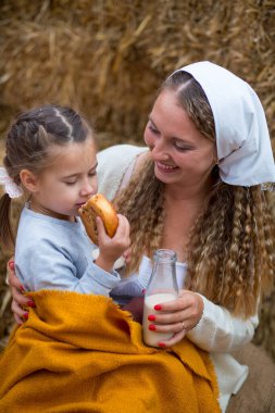 Annesi kızına tarçınlı çörek yediriyor ve arka plandaki saman yığınlarına karşı içmesi için süt veriyor.