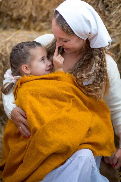 stock image Mom holds in her arms a little daughter with bare feet, wrapped in a yellow cloth, sitting on burlap in stacks of straw and pumpkins and gently hugs